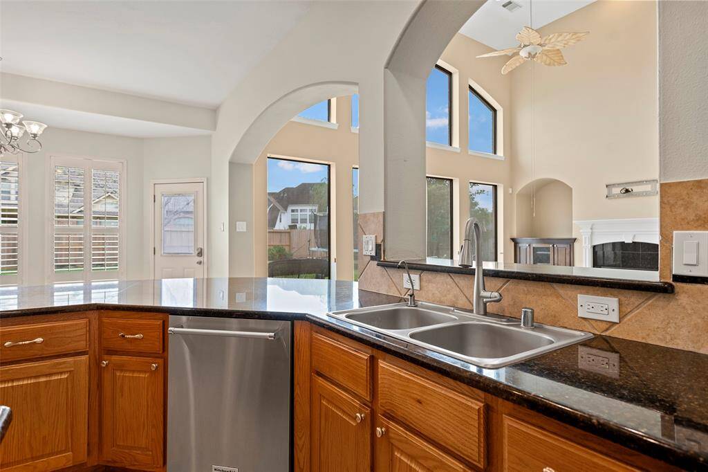 A modern kitchen with granite countertops, a double sink, and stainless-steel dishwasher. Large windows and an open archway offer a view into a bright living area with high ceilings enhancing the spacious feel.