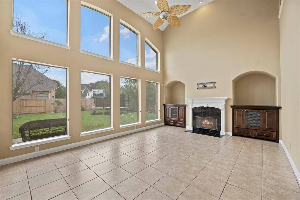 This spacious living room features a high ceiling with large windows that provide ample natural light and a view of the backyard. It has a cozy fireplace framed by elegant built-in cabinets and tiled flooring.