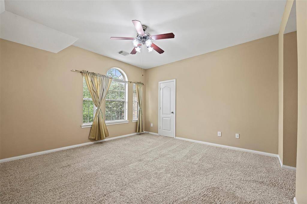 This spacious bedroom features neutral walls, plush carpet, and a ceiling fan with lights. It has a large window with elegant drapes, offering plenty of natural light, and a walk-in closet.