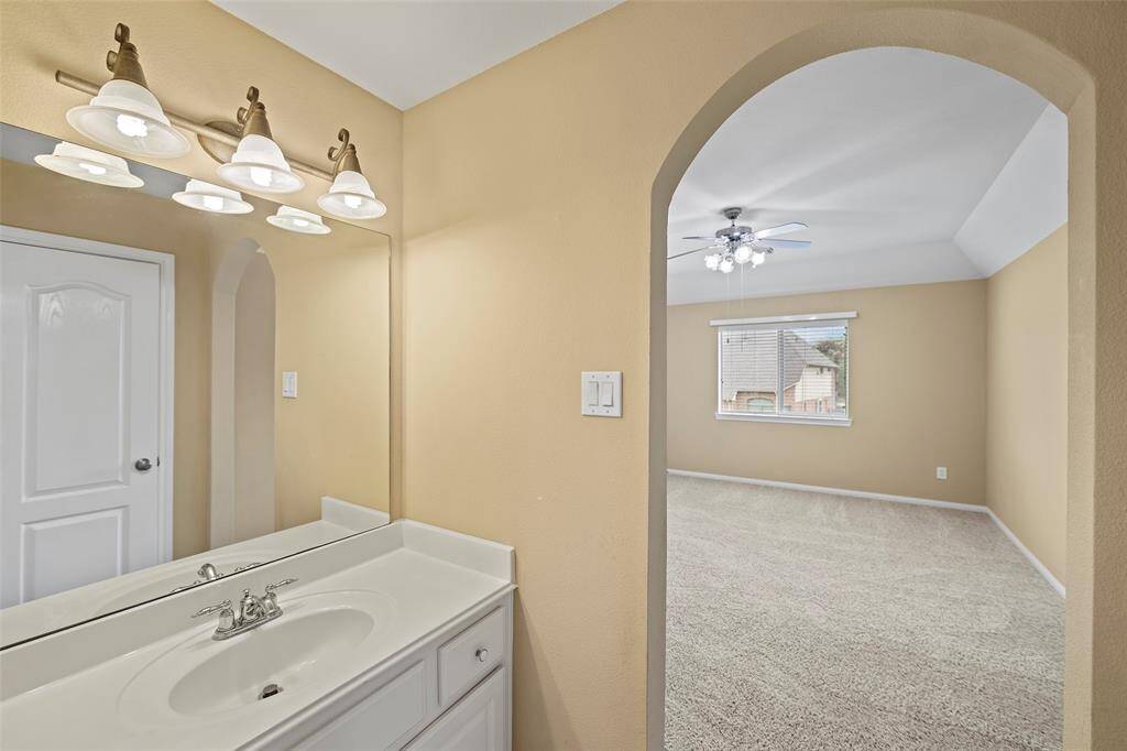 A bright, neutral-toned bathroom with a white vanity and mirror. An arched doorway leads to the adjacent secondary bedroom, creating an open and inviting layout.