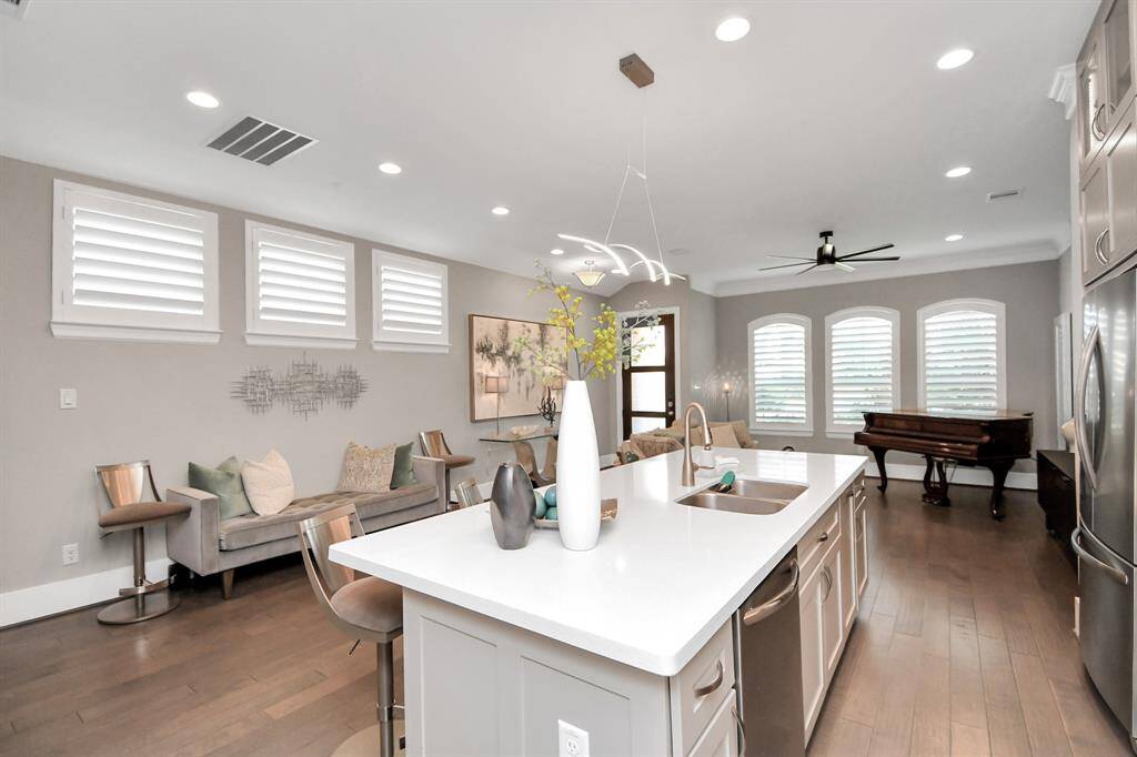 This photo showcases a modern, open-concept living space with an island kitchen, elegant lighting, and hardwood floors. The area features a cozy sitting nook, large windows with shutters, and a grand piano, offering a blend of style and functionality.