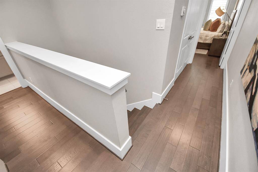 A well-lit hallway with hardwood floors leads to a bedroom. The space features modern white trim and a simple railing along the staircase.