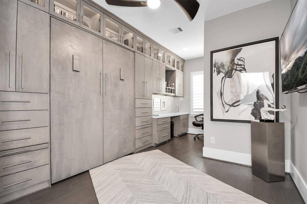 This photo showcases a sleek, modern office space with ample built-in storage and a Murphy bed. There's a stylish desk area with shelving, a large abstract art piece, and a mounted TV. The room has a neutral color palette, dark wood flooring, and a ceiling fan, creating a functional and contemporary work environment.