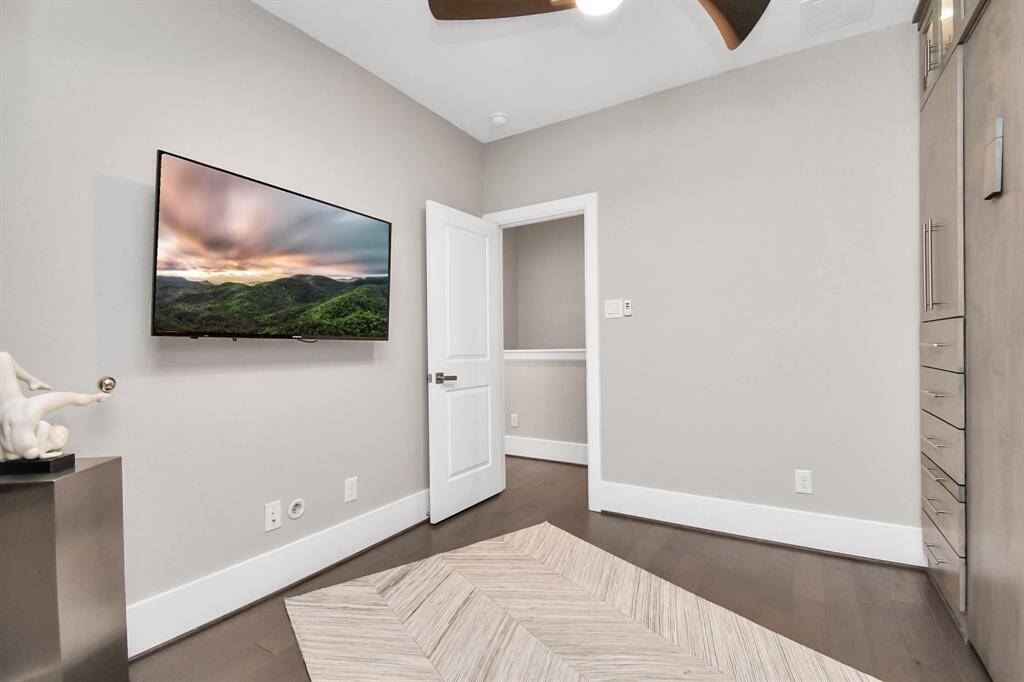 This room features a modern, minimalist design with neutral walls, a mounted TV, and sleek built-in storage. It has dark wood flooring and a stylish ceiling fan, creating a cozy and functional space.