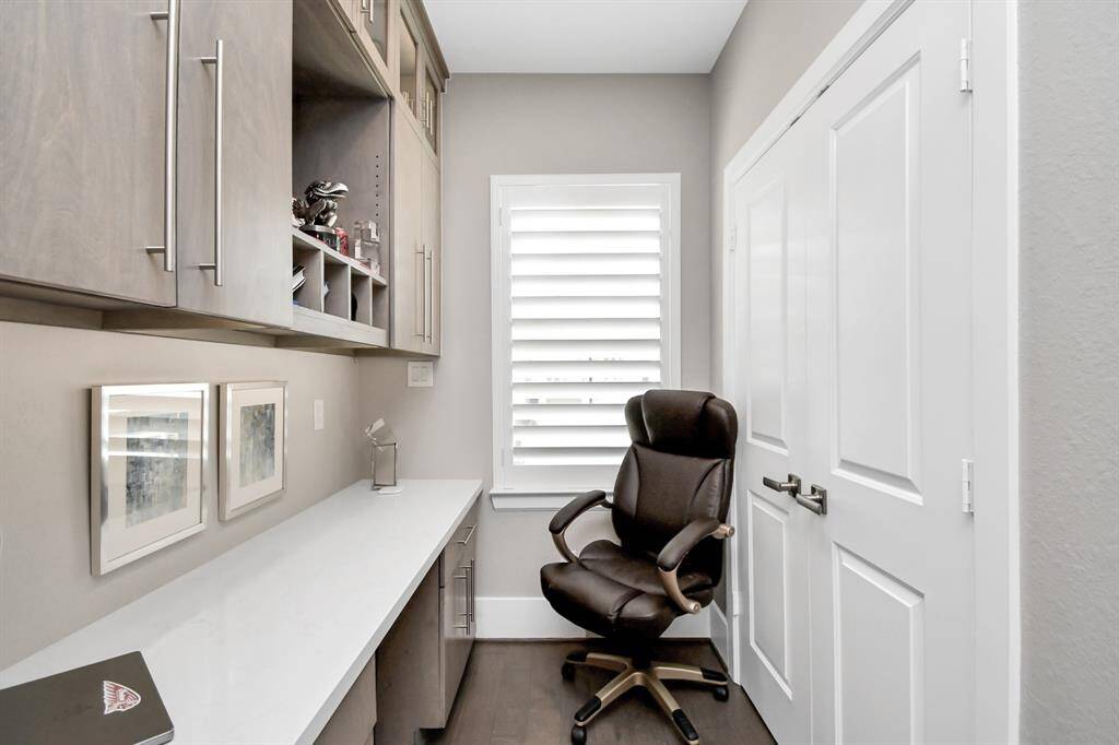 This photo shows a compact home office with built-in cabinets, a sleek white desk, and a comfortable swivel chair. A window with shutters provides natural light, and the space is accented with framed artwork.