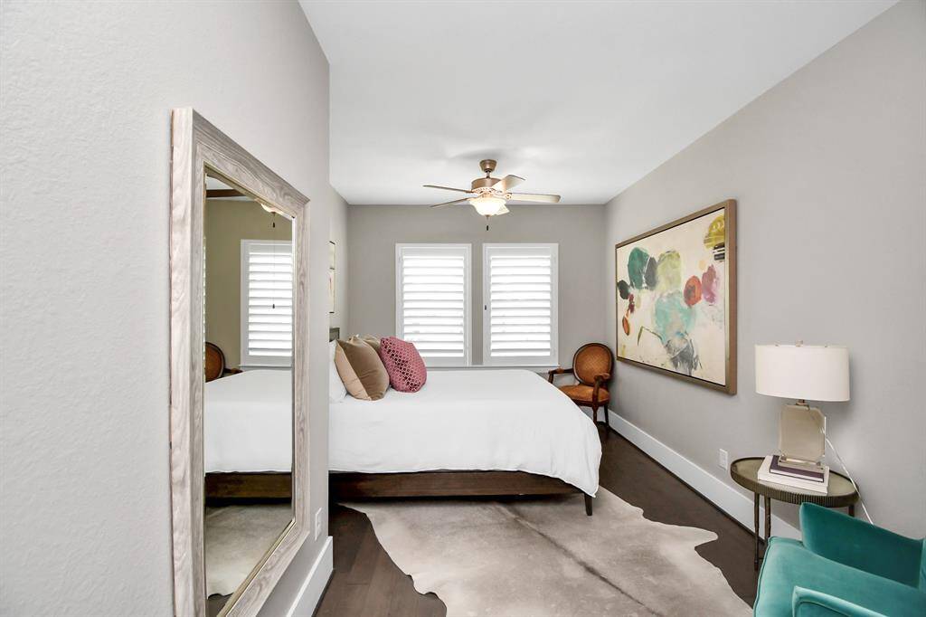 This cozy bedroom features a modern design with soft gray walls, a ceiling fan, and dark hardwood floors. It includes a large wall mirror, stylish abstract art, and ample natural light from two windows with shutters. The room is furnished with a comfortable bed, elegant chair, and a side table with a lamp.