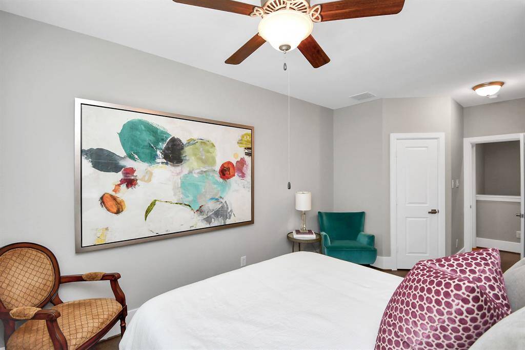 This bedroom features neutral walls and a ceiling fan, with modern abstract art adding a splash of color. It includes a cozy sitting area with a teal chair, a classic wooden chair, and ample natural light.