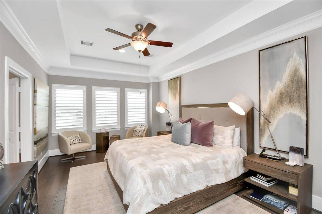 This bedroom features a modern design with a tray ceiling and a ceiling fan. It includes a comfortable bed with plush pillows, two contemporary bedside lamps, and stylish artwork. Natural light fills the room through three large windows with shutters. A cozy seating area with two chairs sits by the windows, complemented by dark wood flooring and neutral wall colors.