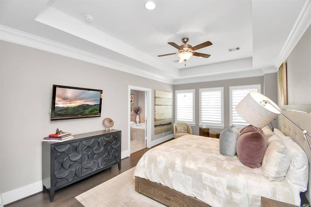 This spacious bedroom features a cozy bed with plush pillows, a ceiling fan, and natural light from large windows with shutters. It includes a wall-mounted TV, stylish dresser, and neutral decor, offering a relaxing and modern atmosphere.