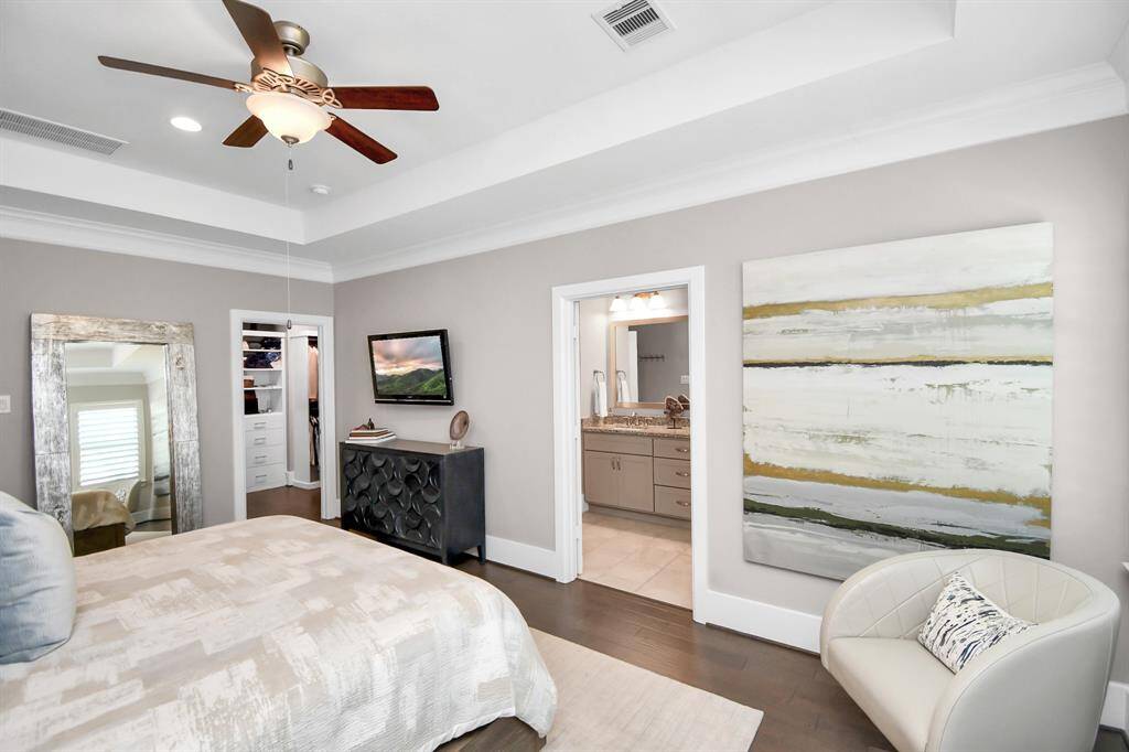 This bedroom features soft neutral tones, a ceiling fan, and modern decor. There's a large abstract painting, a cozy chair, and a stylish dresser. The room offers a view into a spacious walk-in closet and an attached bathroom with double sinks.