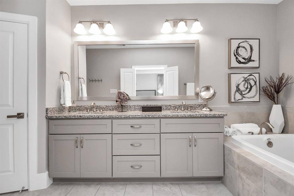 This bathroom features a modern double vanity with granite countertops, framed by a large mirror and elegant lighting. Adjacent is a tiled bathtub, adding a touch of luxury. Neutral tones and contemporary decor create a serene atmosphere.