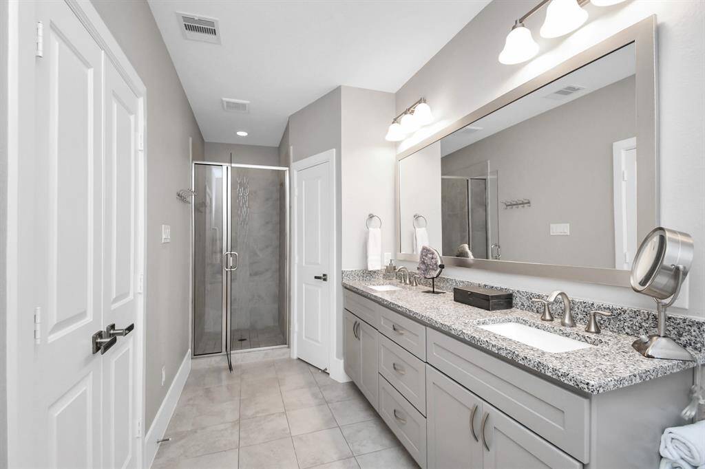 This modern bathroom features a sleek dual-sink vanity with granite countertops, ample storage, and a large mirror. It includes a spacious glass-enclosed shower and neutral-colored tiles for a clean, elegant look.