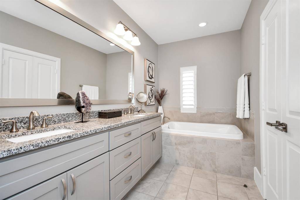 This modern bathroom features a double-sink vanity with granite countertops, a large mirror, a soaking tub, and neutral tile flooring. The space is well-lit and has a clean, contemporary design.