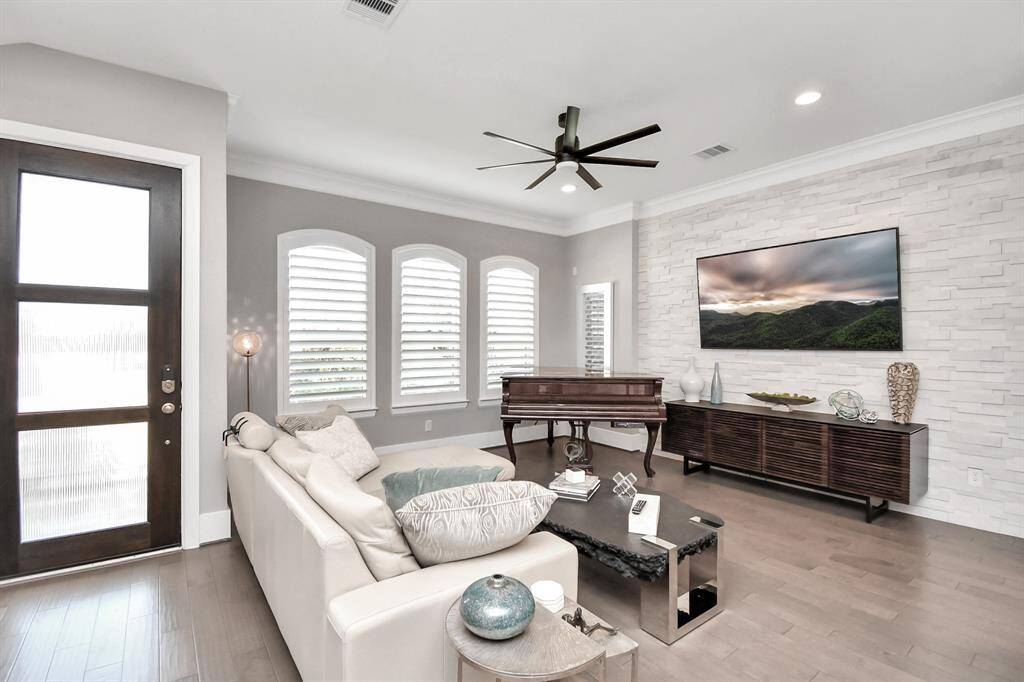This modern living room features a cozy layout with a sleek white sofa, a large wall-mounted TV, and a stylish ceiling fan. The space is brightened by multiple arched windows with shutters and a textured feature wall. A grand piano adds an elegant touch, and there's ample space for entertaining or relaxing.
