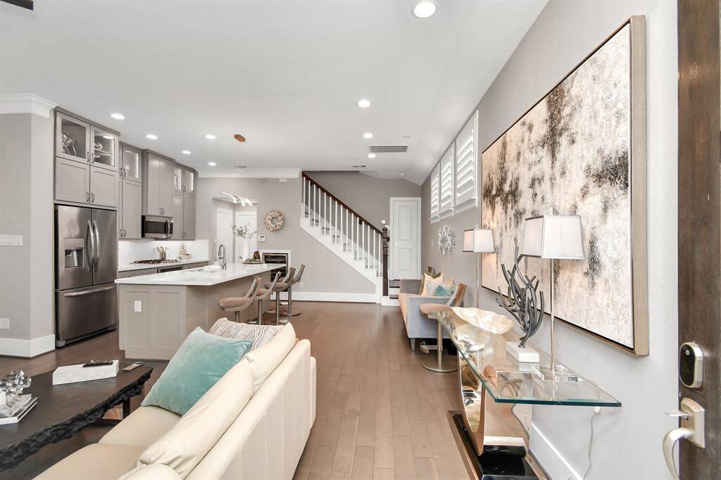 This photo showcases a modern open-concept living area with a sleek kitchen featuring stainless steel appliances and a large island. The space includes a cozy seating area, contemporary artwork, and stylish decor, leading to a staircase with clean lines and abundant natural light.