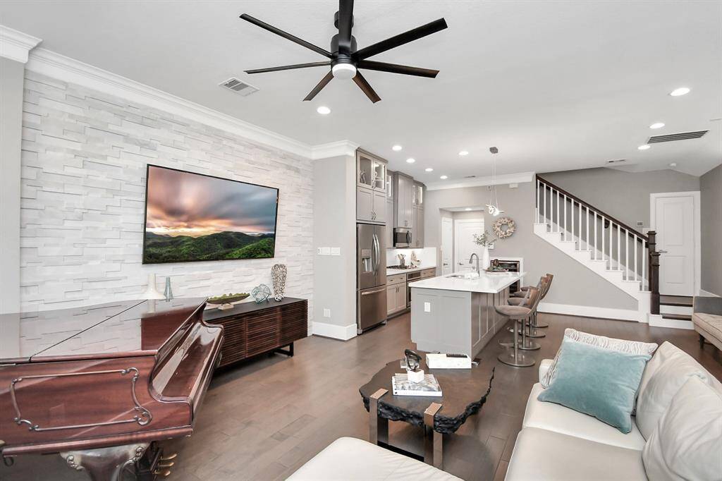 This modern open-concept living space features a sleek kitchen with stainless steel appliances, an island with bar seating, and a cozy living area. The room is enhanced by a stone accent wall with a mounted TV, elegant dark wood flooring, and stylish furnishings. A staircase leads to the upper level, adding to the inviting and functional layout.