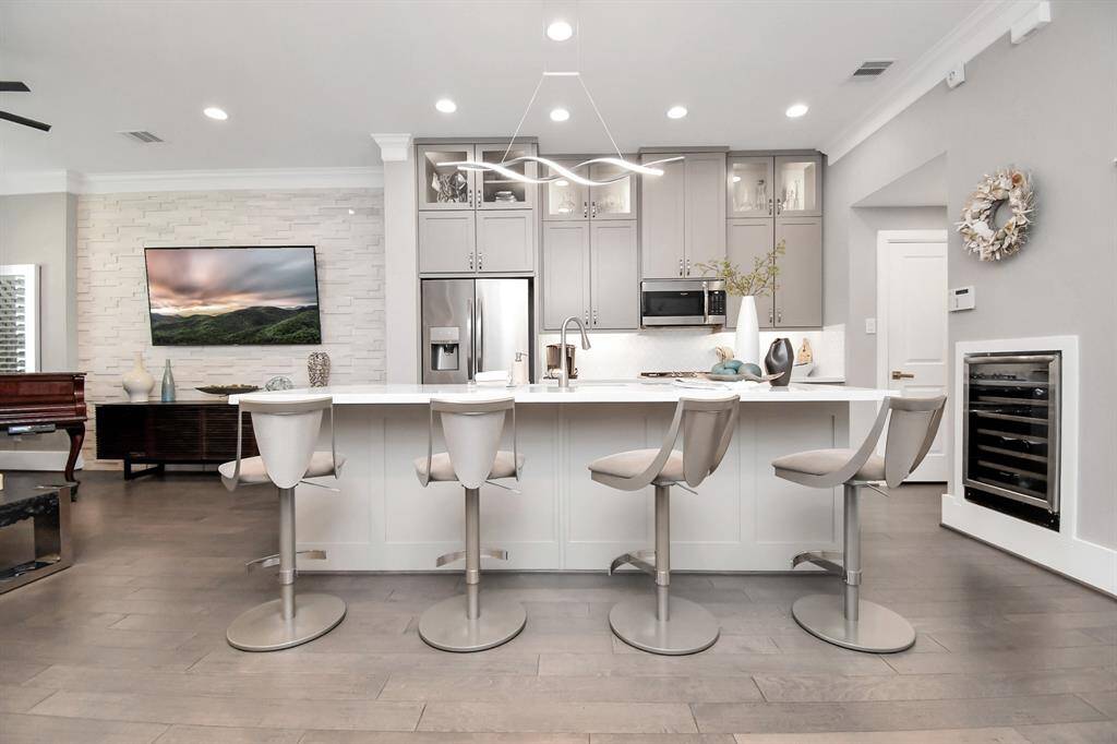 This modern kitchen features sleek gray cabinetry and stainless steel appliances. A large island with stylish bar stools provides ample seating and workspace. The open layout flows into a living area with a wall-mounted TV and tasteful decor, creating a welcoming and functional space.