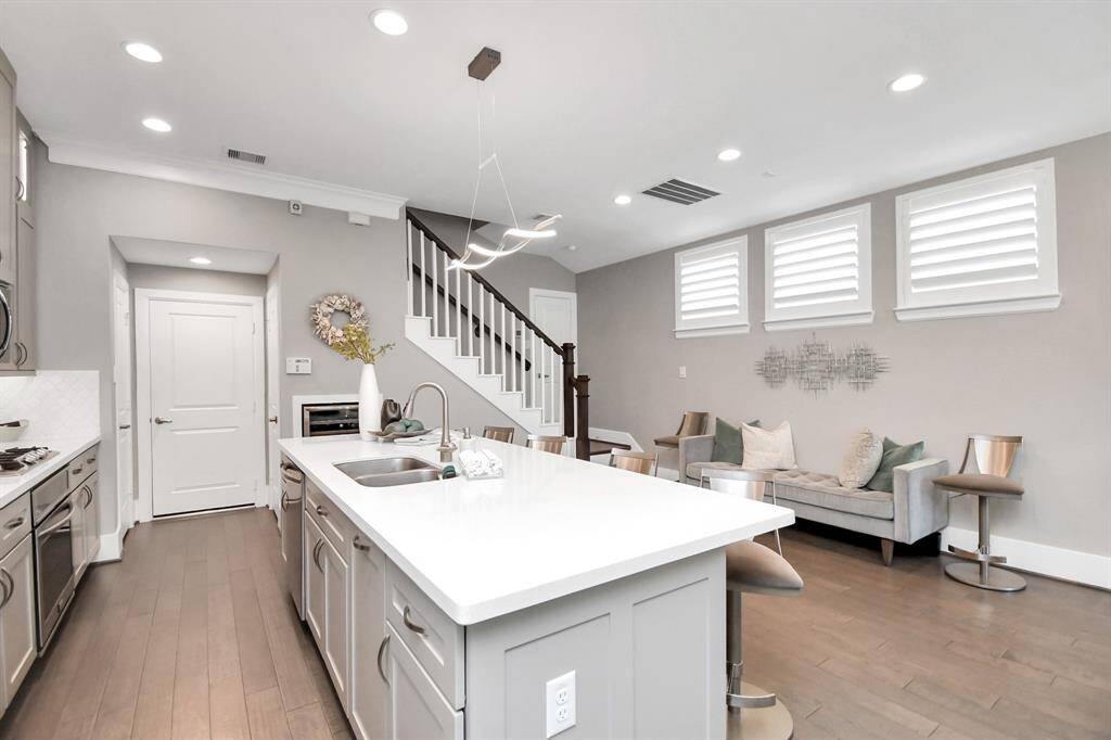 This modern open-concept kitchen and living area features a sleek design with hardwood floors, a large island with a sink, stainless steel appliances, and ample natural light from three windows. The space is inviting, with contemporary furniture and stylish lighting, perfect for entertaining or relaxing.
