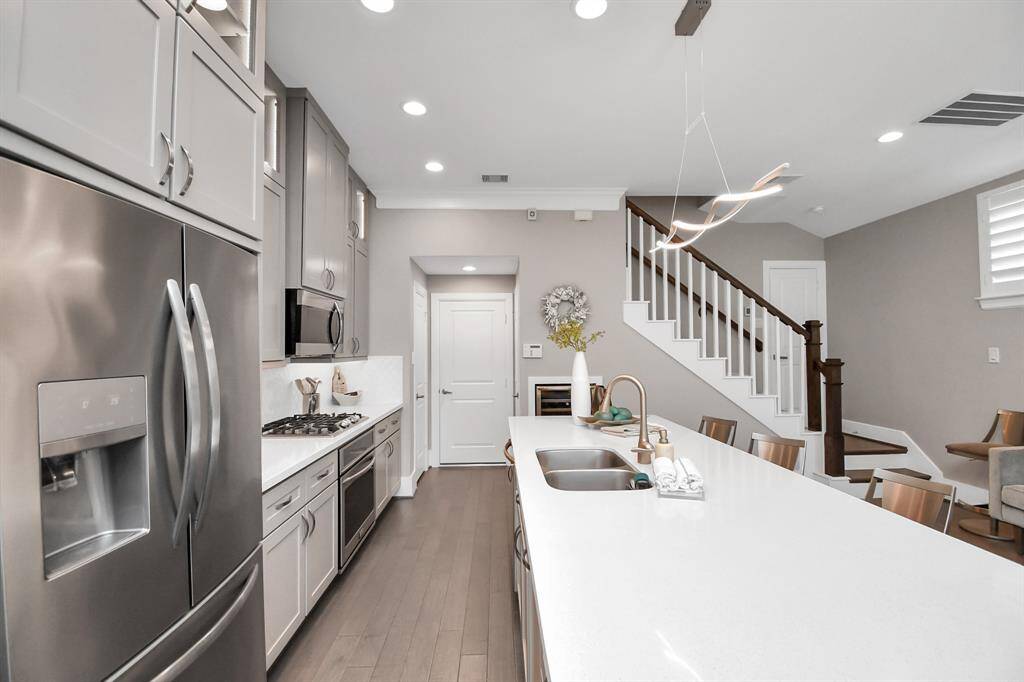 This modern kitchen features sleek stainless steel appliances, a spacious island with a double sink, and elegant cabinetry. The open layout flows into the living area, complemented by a stylish staircase and contemporary lighting. Ideal for both cooking and entertaining.