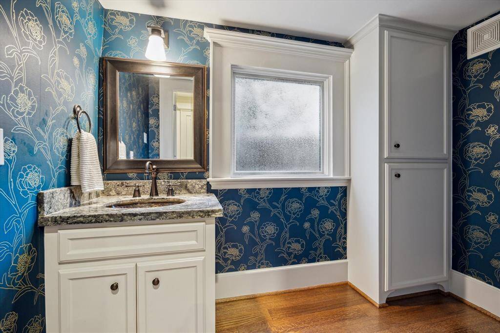 Off the family room, this charming, windowed powder room features designer wallpaper, ample storage, and a granite-topped vanity with oil-rubbed bronze hardware.