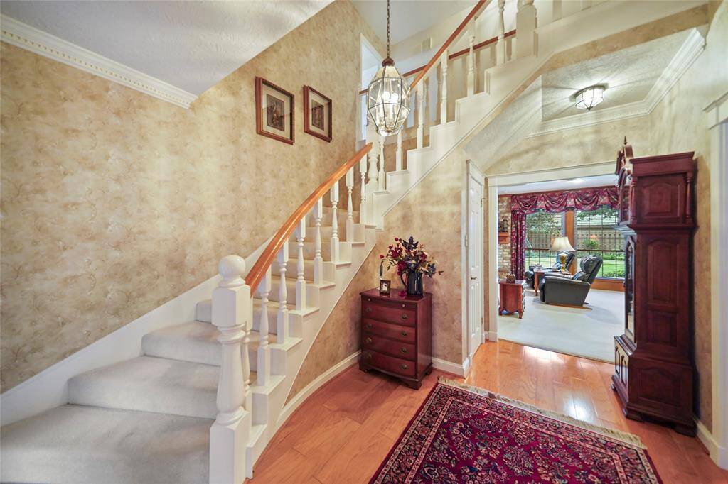 Welcoming entryway with staircase and heavy wooden banister. A cozy family room is visible through an arched doorway featuring large windows.