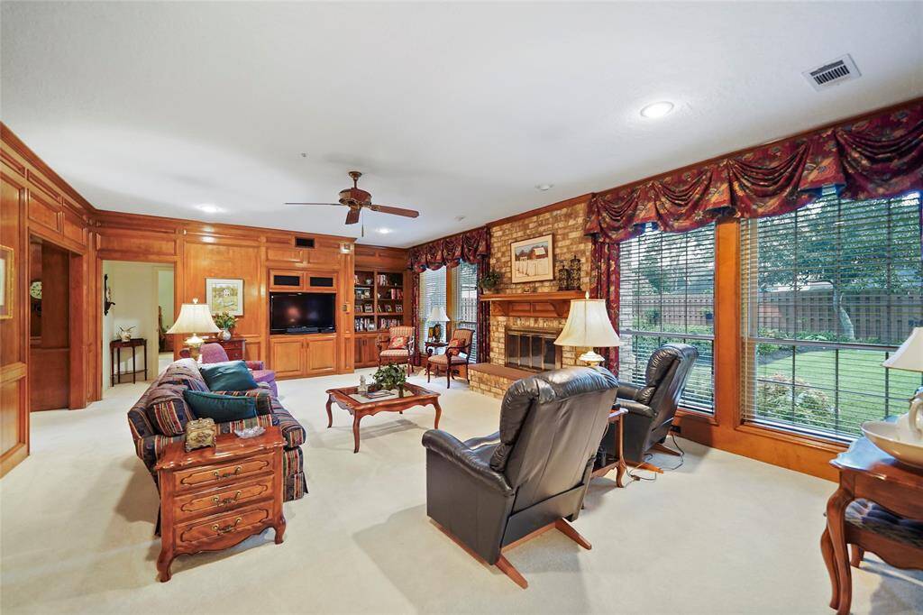 Spacious living room with warm wood paneling, featuring a cozy brick fireplace and large windows offering views of the landscaped yard. The room includes built-in shelving and a welcoming and comfortable atmosphere.