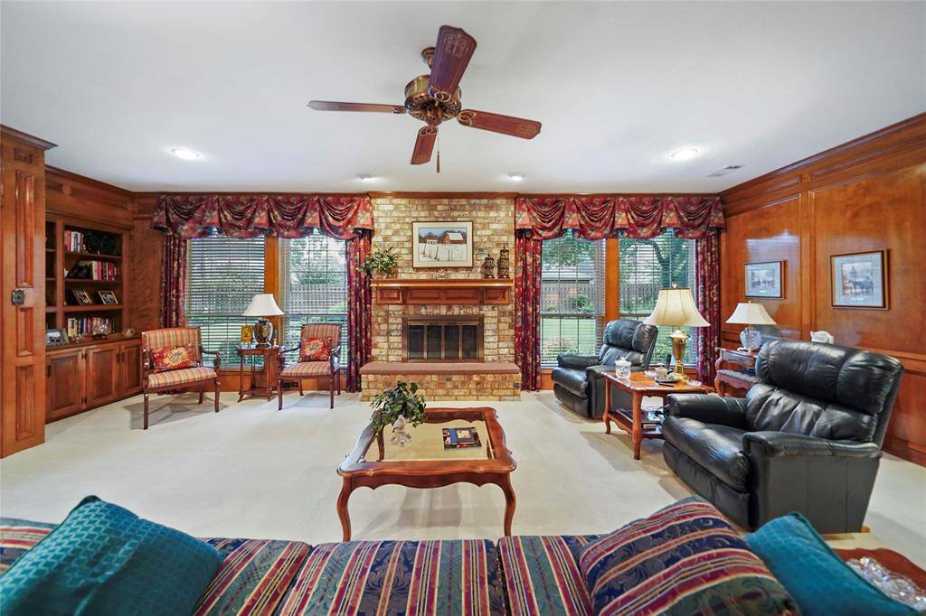 This inviting living room features warm wood paneling, a brick fireplace, and large windows overlooking the massive backyard. Built-in bookshelves and stylish ceiling fan add to its cozy charm.