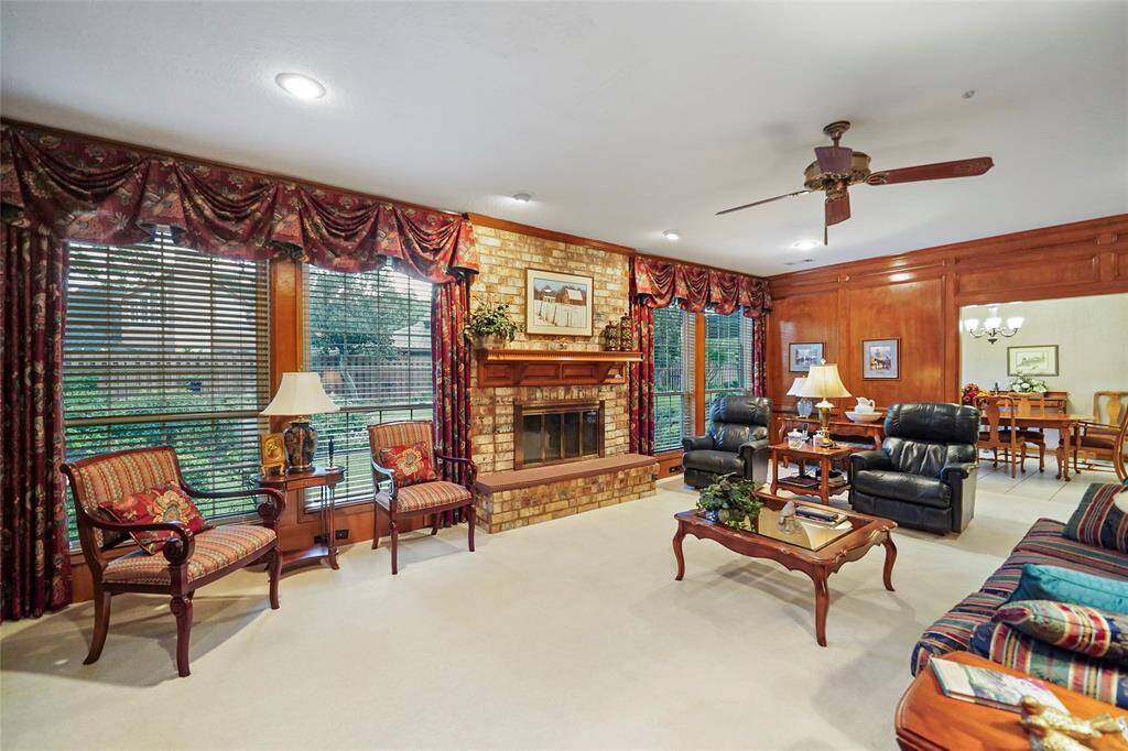 This cozy living room features a warm brick fireplace, elegant wood paneling, and large windows with inviting atmosphere. The space opens to a dining area breakfast area/kitchen, perfect for entertaining.