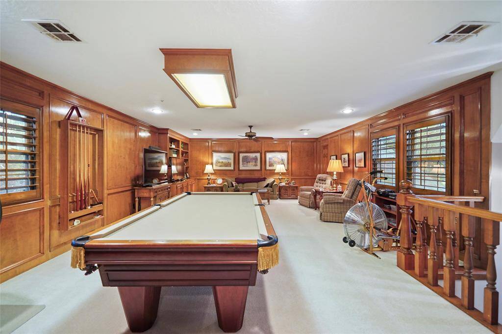 Spacious game room with wood paneling, featuring a pool table, built-in shelving, and cozy seating area. It has ample lighting and a window with shutters, creating a warm and inviting atmosphere.