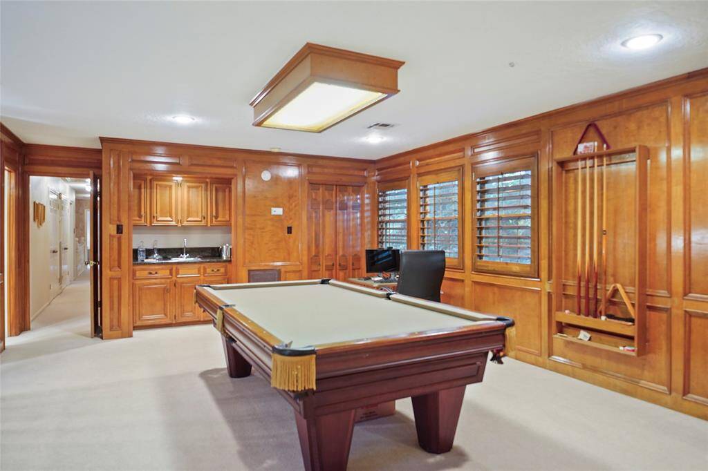 This room features rich wood paneling, a built-in bar with a sink, and a pool table as the centerpiece. Shuttered windows provide natural light, complemented by recessed ceiling lights. Ideal for entertainment and relaxation.