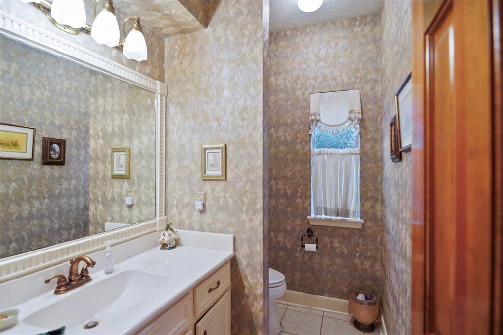 Half-bath features a classic design with textured wallpaper, a large mirror, and an elegant vanity with brass fixtures. It has a window with a decorative curtain, providing natural light and a cozy touch.