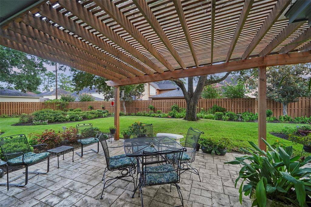 This photo showcases a charming backyard with a covered patio featuring a wooden pergola and outdoor seating area. The lush, well-maintained garden includes a variety of plants and a large tree, offering a private and inviting outdoor space.