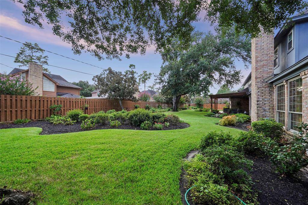 Beautifully landscaped backyard with lush green grass, mature trees, and well-maintained flower beds. The double-lot backyard space is bordered by a well-maintained fence, providing privacy, and features a charming stone patio area attached to the house. Ideal for outdoor relaxation and entertaining.