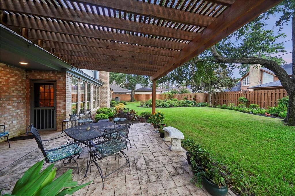 This photo showcases a charming backyard with a covered patio featuring a pergola, stone patterned decking, and outdoor seating area. The well-maintained lawn is bordered by a wooden fence and lush greenery, creating a private and inviting outdoor space.