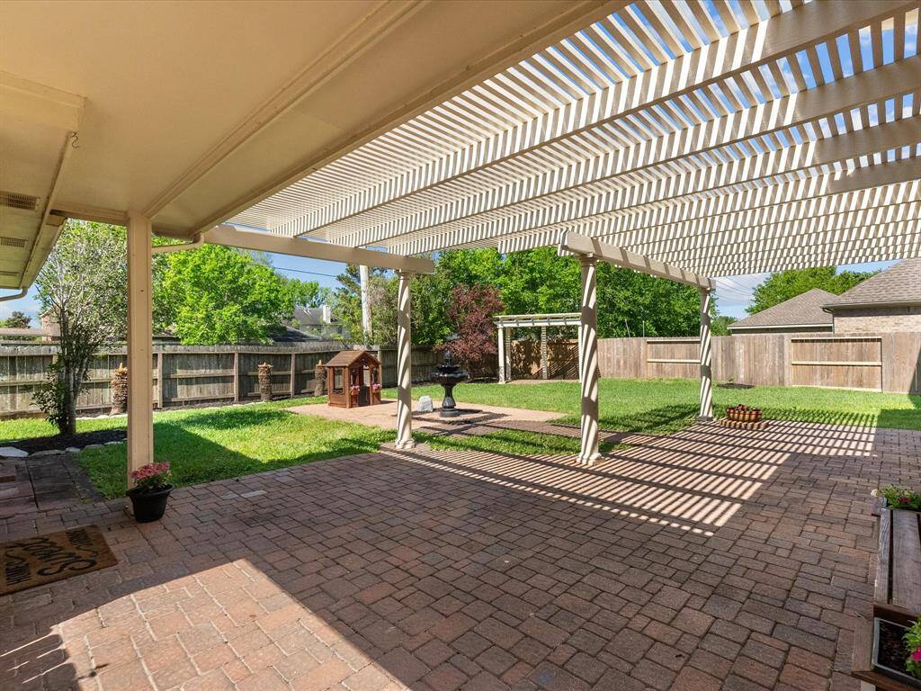 Spacious backyard with a brick patio and a large pergola providing shade. Features a grassy area, a small playhouse, and a fountain, all enclosed by a wooden fence, offering a private and inviting outdoor space.