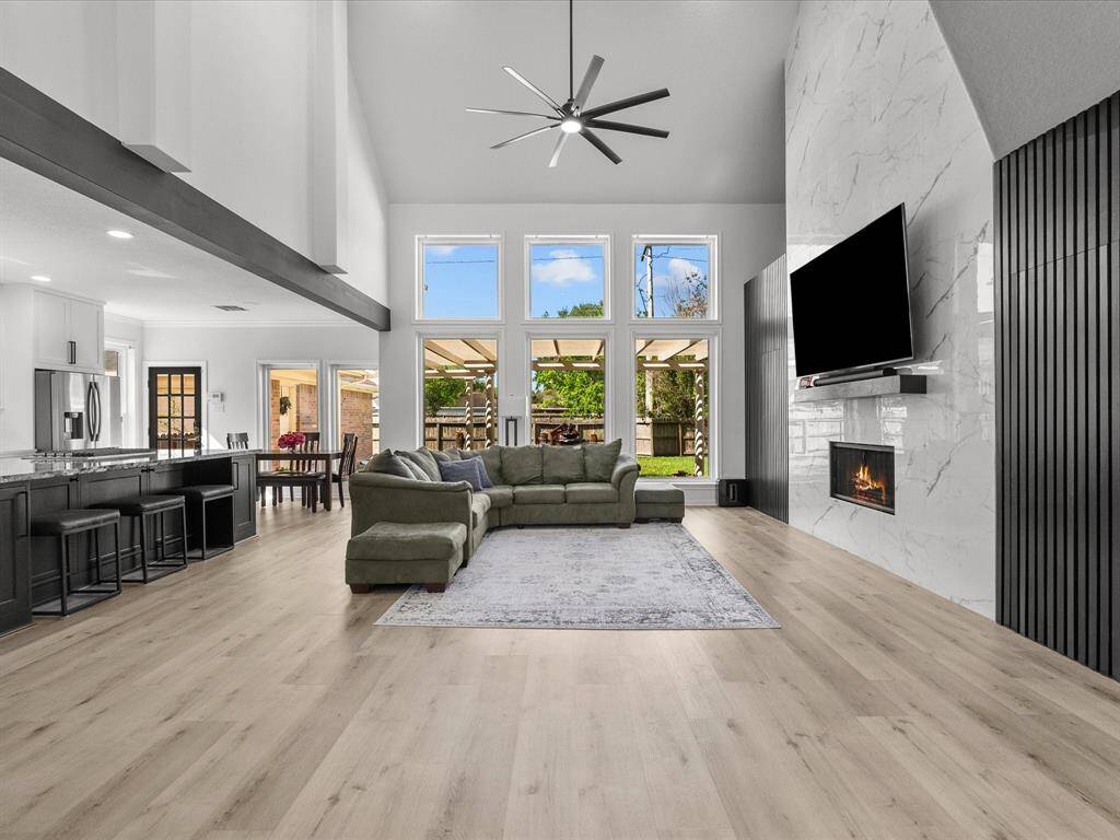 This spacious living area features a high ceiling, large windows, and a modern open-plan design. It includes a cozy fireplace, stylish flooring, and a seamless flow into the kitchen and dining area. Natural light fills the room, offering a view of the outdoor patio and garden.
