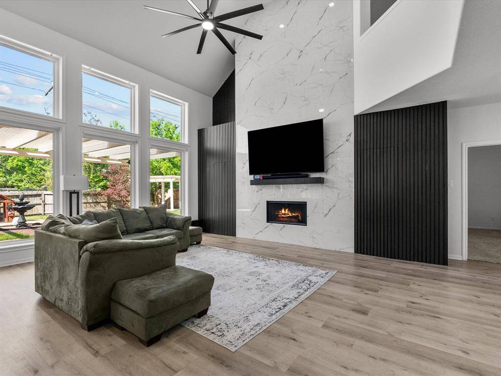 This spacious living room features tall ceilings, large windows for abundant natural light, and a modern marble accent wall with a sleek fireplace. The open layout and elegant wood flooring create a welcoming and contemporary atmosphere along with the accent walls hidden closets