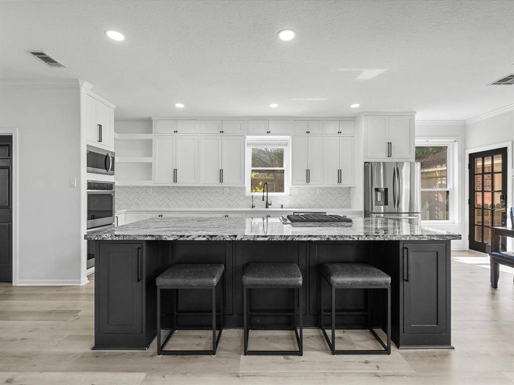 This modern kitchen features a large island with seating, sleek black and white cabinetry, and stainless steel appliances. It has ample natural light and a stylish backsplash, creating a bright and inviting space.