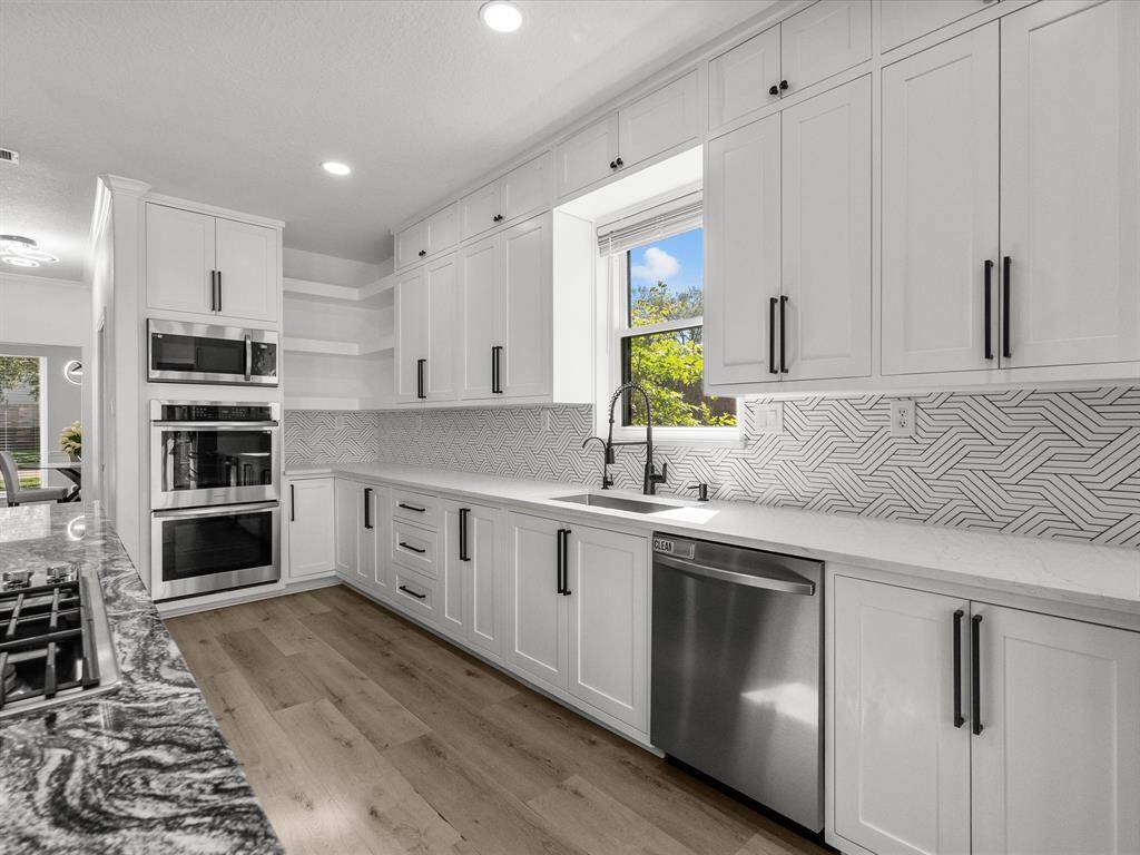 This modern kitchen features sleek white cabinetry with black hardware, a stylish geometric backsplash, and stainless steel appliances. It has ample counter space and natural light from a window above the sink, creating a bright and inviting atmosphere.