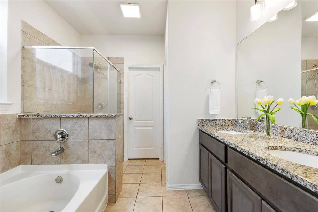 This stylish primary bathroom features dual sinks, rich stained cabinetry with granite countertops, a walk-in shower, and a relaxing soaker tub.