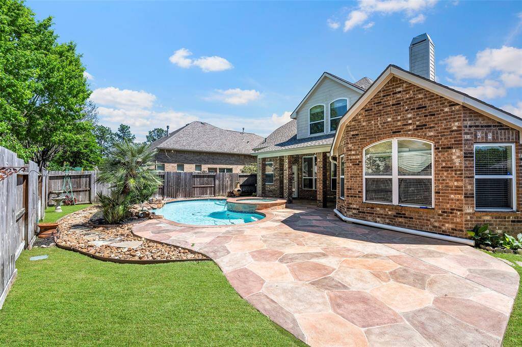 This backyard features a stunning pool with a connected spa, surrounded by a stone patio. The area is landscaped with lush greenery and a wooden fence provides privacy, creating a perfect outdoor retreat.