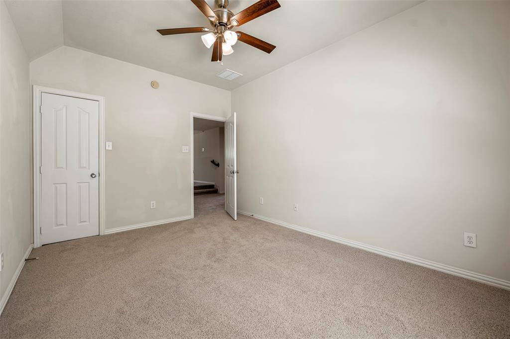 This inviting bedroom boasts neutral walls and carpet, a ceiling fan with lights, and a convenient closet. An open doorway seamlessly connects to a hallway with stairs.
