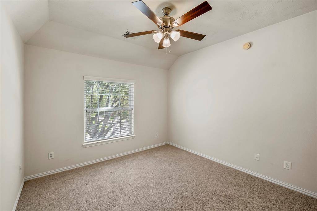 This bedroom features neutral-toned walls and carpet, a ceiling fan with lights, and a closet. It has an open doorway connecting to a hallway with stairs.