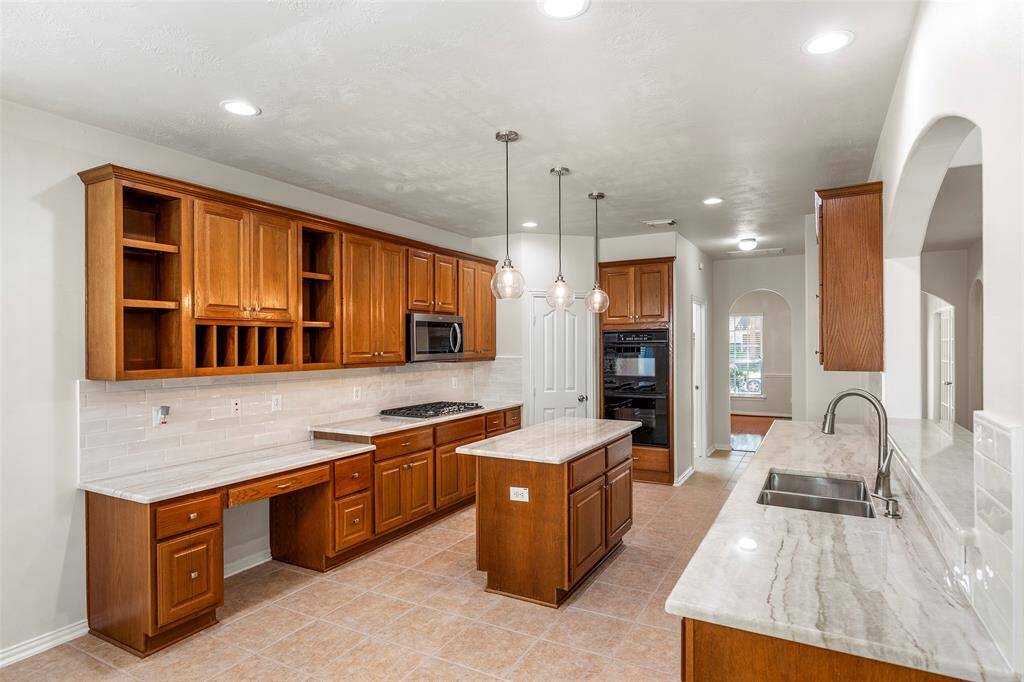 This kitchen features warm wooden cabinetry, modern stainless steel appliances, and a central island with pendant lighting. The tile flooring and neutral backsplash add a stylish touch, while the open layout connects to a sunlit hallway.