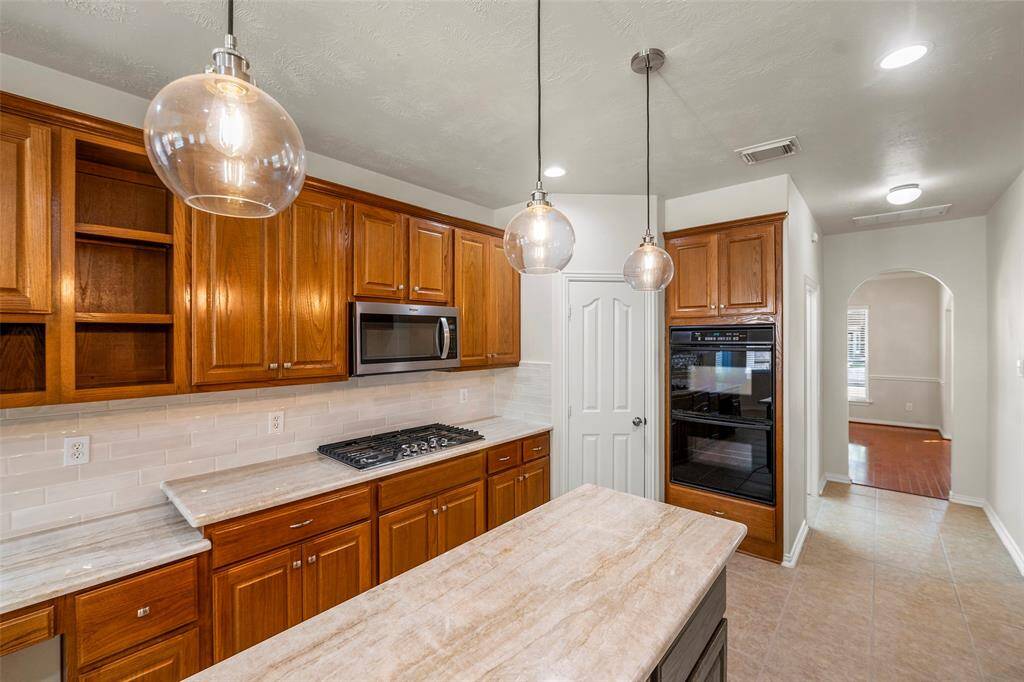 This bright kitchen features a spacious dining area with large windows, tile flooring, and a modern light fixture. It includes ample wooden cabinetry, a marble countertop, and a door leading to an outdoor space.