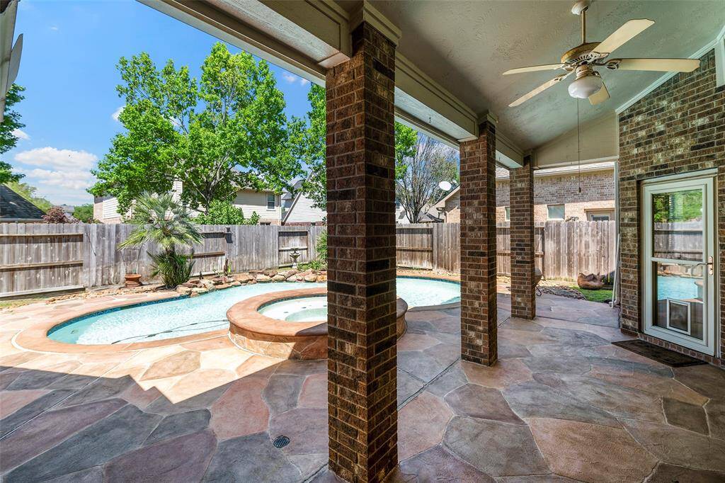 A covered patio with brick columns and a ceiling fan is showcased leading to a backyard featuring a pool with a stone border. The area is enclosed by a wooden privacy fence, with mature trees providing natural shade.