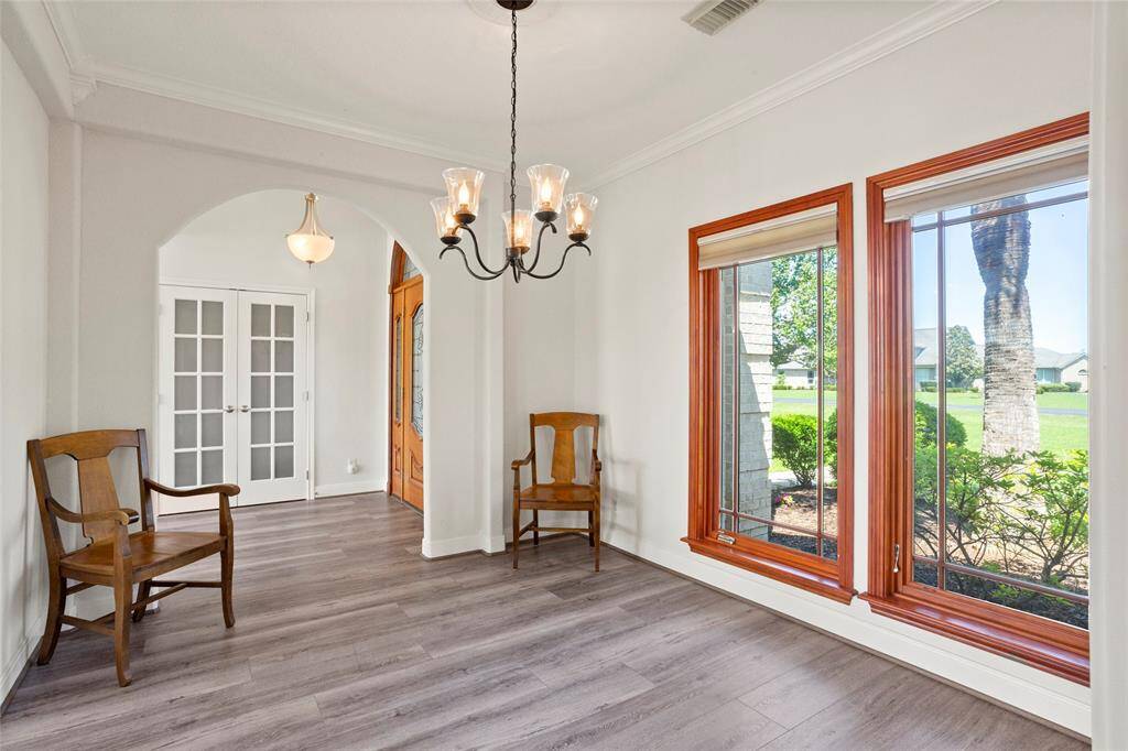 Bright, inviting dining room with large windows, wood LVP flooring, and a charming chandelier. Features arched doorway and French doors, offering a warm, classic feel with views of a lush green lawn.
