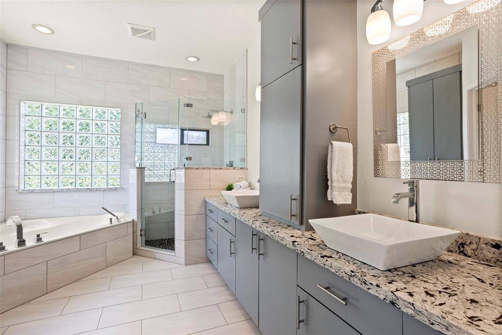 The updated primary bathroom features a spacious layout with a glass-enclosed shower, large jetted tub, and dual square sinks on a sleek granite countertop. The room is bright with stylish tile flooring and ample storage in contemporary cabinetry.