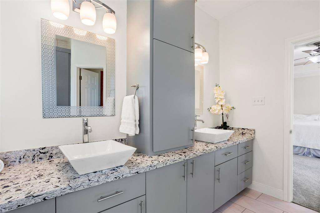 The primary bathroom features a modern design with dual sinks, sleek grey cabinetry, and a granite countertop. It has stylish lighting fixtures and a decorative mirror, creating a bright and elegant space.