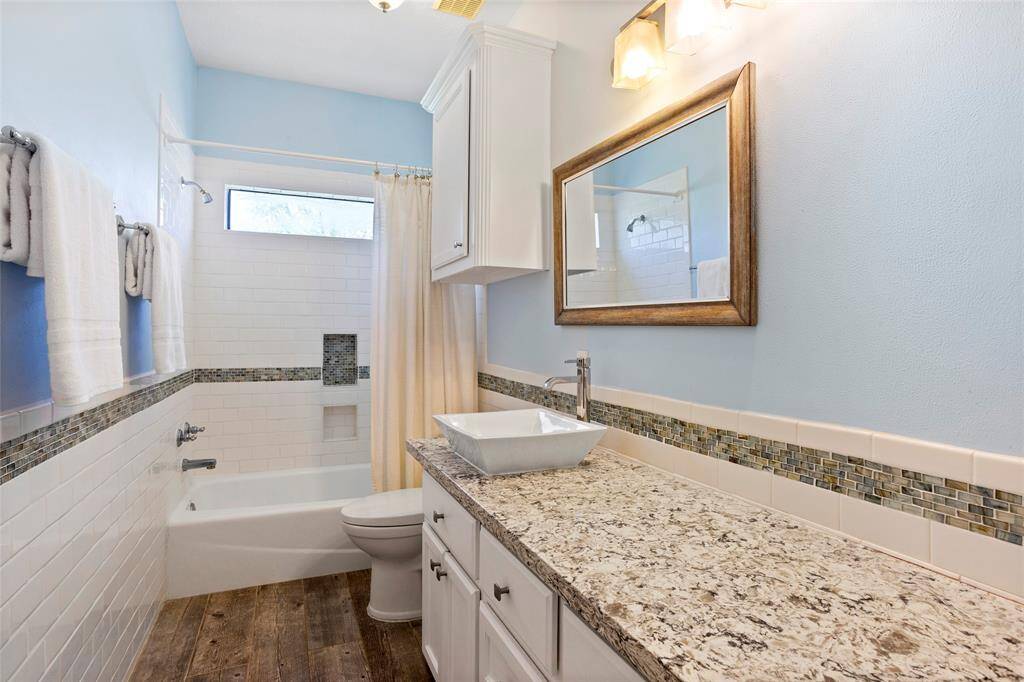 This downstairs bathroom features a modern design with a granite countertop, above-counter sink, and wood-framed mirror. It includes a tub with a custom tiled backsplash, wood-look tile flooring, and soft blue walls for a serene atmosphere.